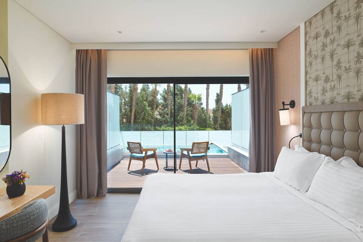 A modern hotel room with a large bed, a view of a pool, and two chairs by the window.