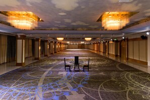 A long, narrow hotel corridor with chandeliers, a patterned carpet, and a central table with chairs.