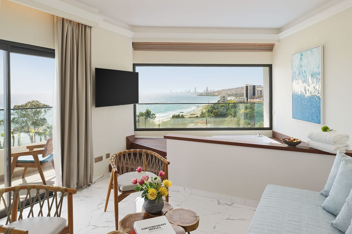 A modern hotel room with a large balcony, a flat-screen TV, a comfortable seating area, and a view of the sea and city skyline.