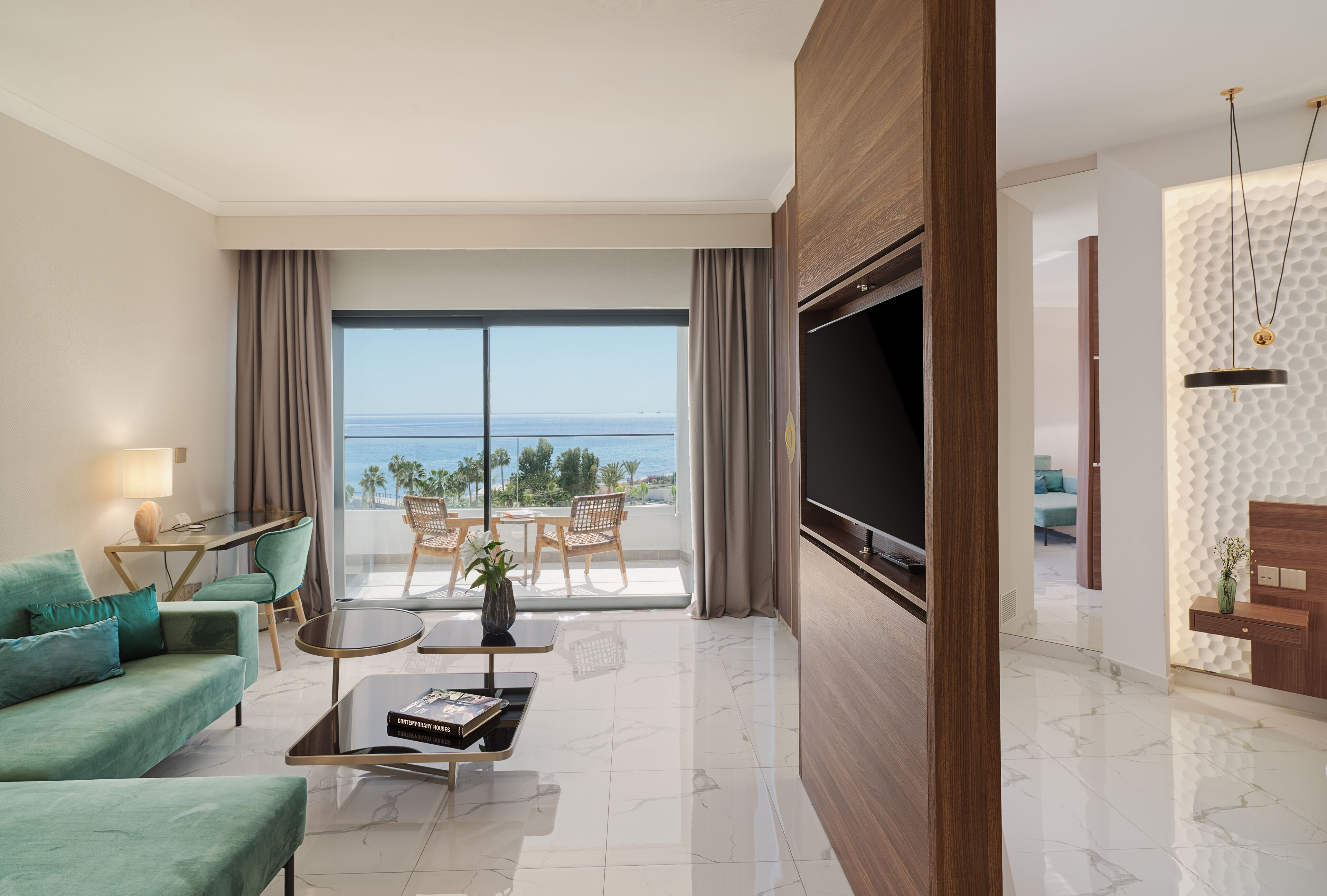 Habitación de hotel moderna con un televisor grande, un sofá, una mesa de centro y un balcón con vistas al mar.