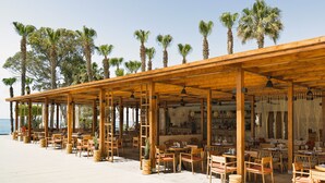 Ein Straßencafé mit Holztischen und -stühlen, Palmen und Blick auf das Meer.