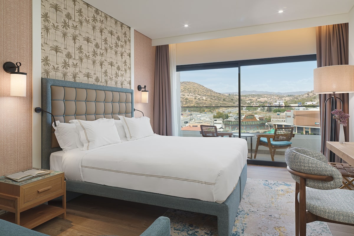 A hotel room with a large bed, a desk, a chair, and a view of the city.