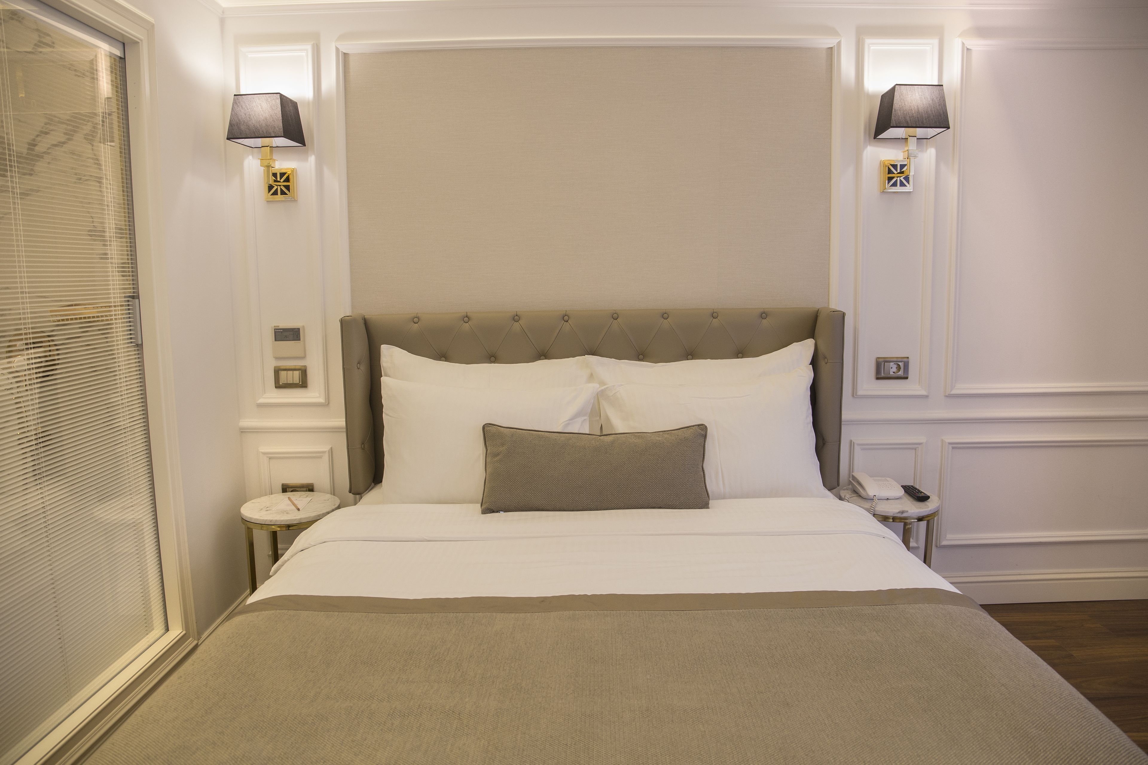 A neatly made bed with a headboard, two bedside lamps, a bedside table with a phone, and a window with blinds.