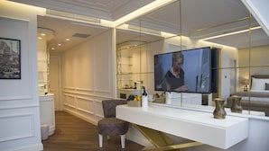Una habitación de hotel moderna con una mesa blanca tipo consola, un televisor y una zona de estar con un taburete y una silla estampada.
