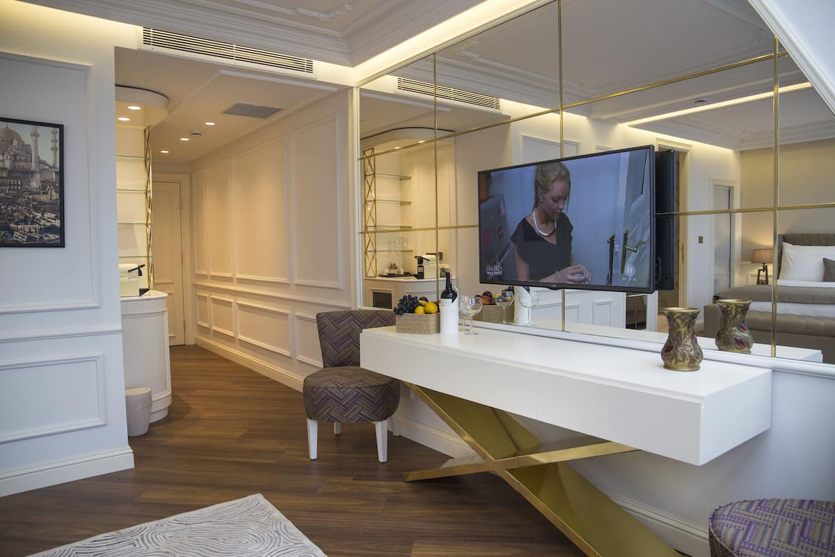 Una habitación de hotel moderna con una mesa blanca tipo consola, un televisor y una zona de estar con un taburete y una silla estampada.