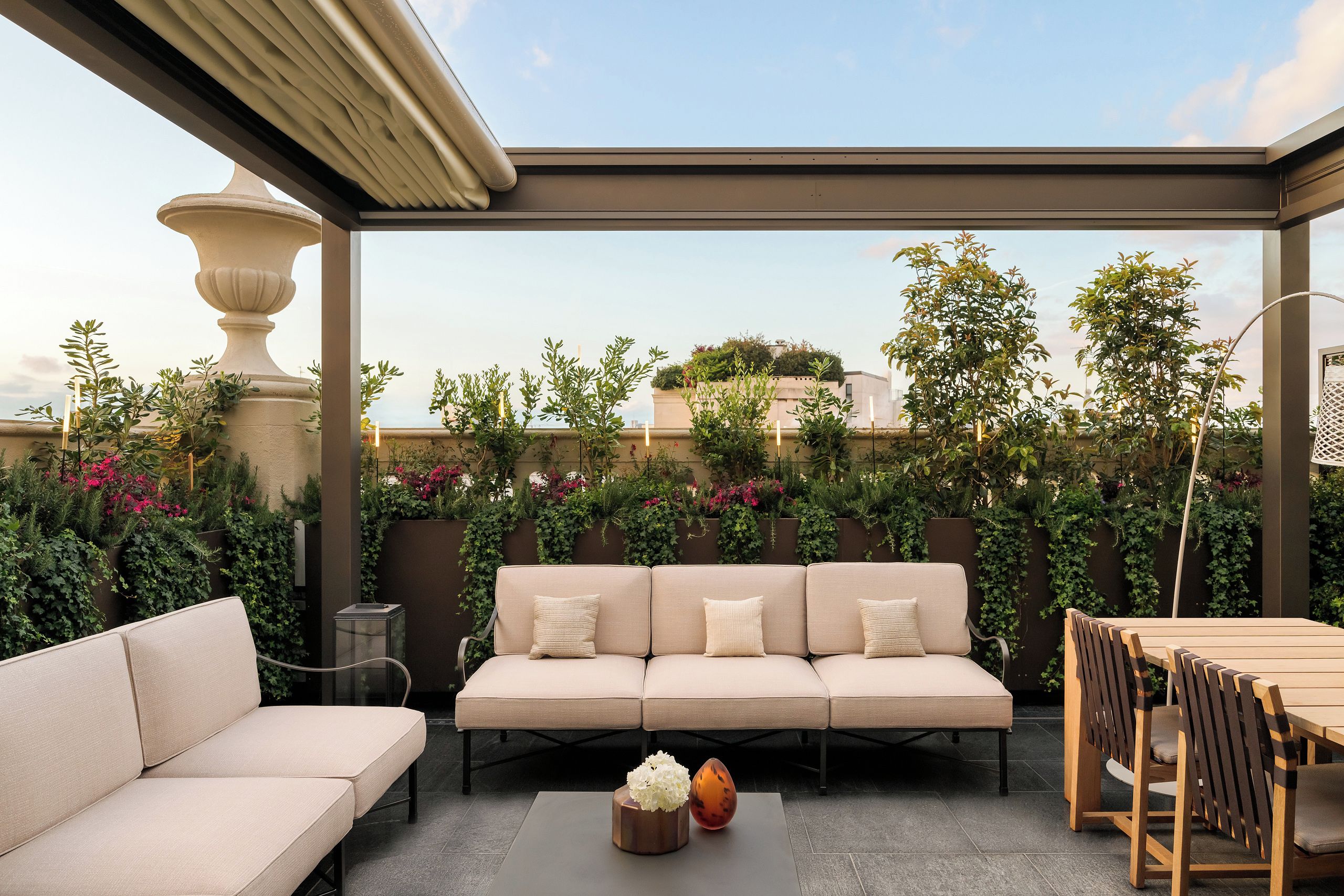 Eine Dachterrasse mit einem beigen Sofa, einem Holzesstisch und Stühlen. Es befinden sich Blumentöpfe und eine Zierurne dort.