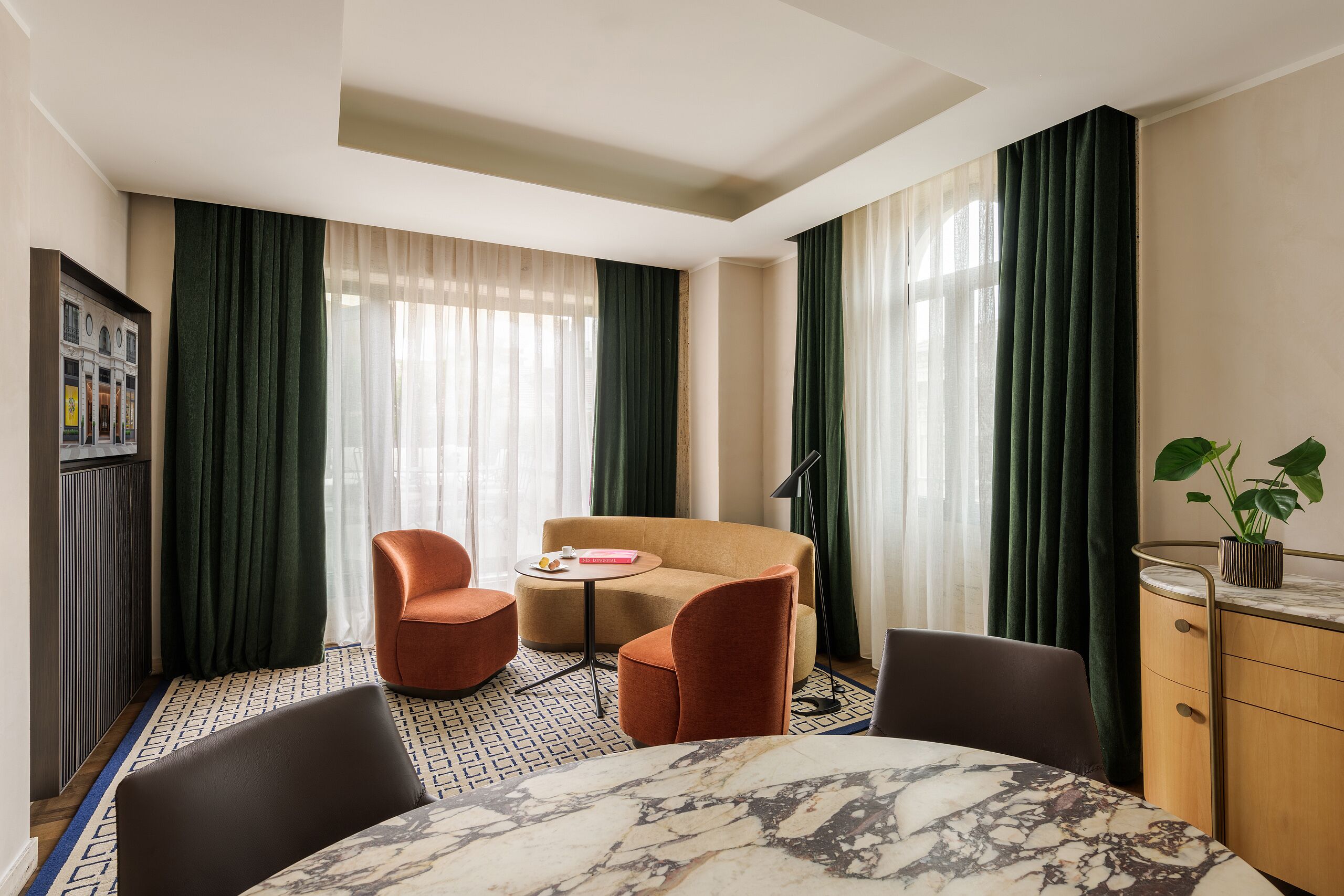 Ein Hotelzimmer mit einem Esstisch aus Marmor, zwei Sesseln, einer Couch, einem kleinen runden Tisch mit Buch und Lampe sowie einem Schrank mit Pflanze.
