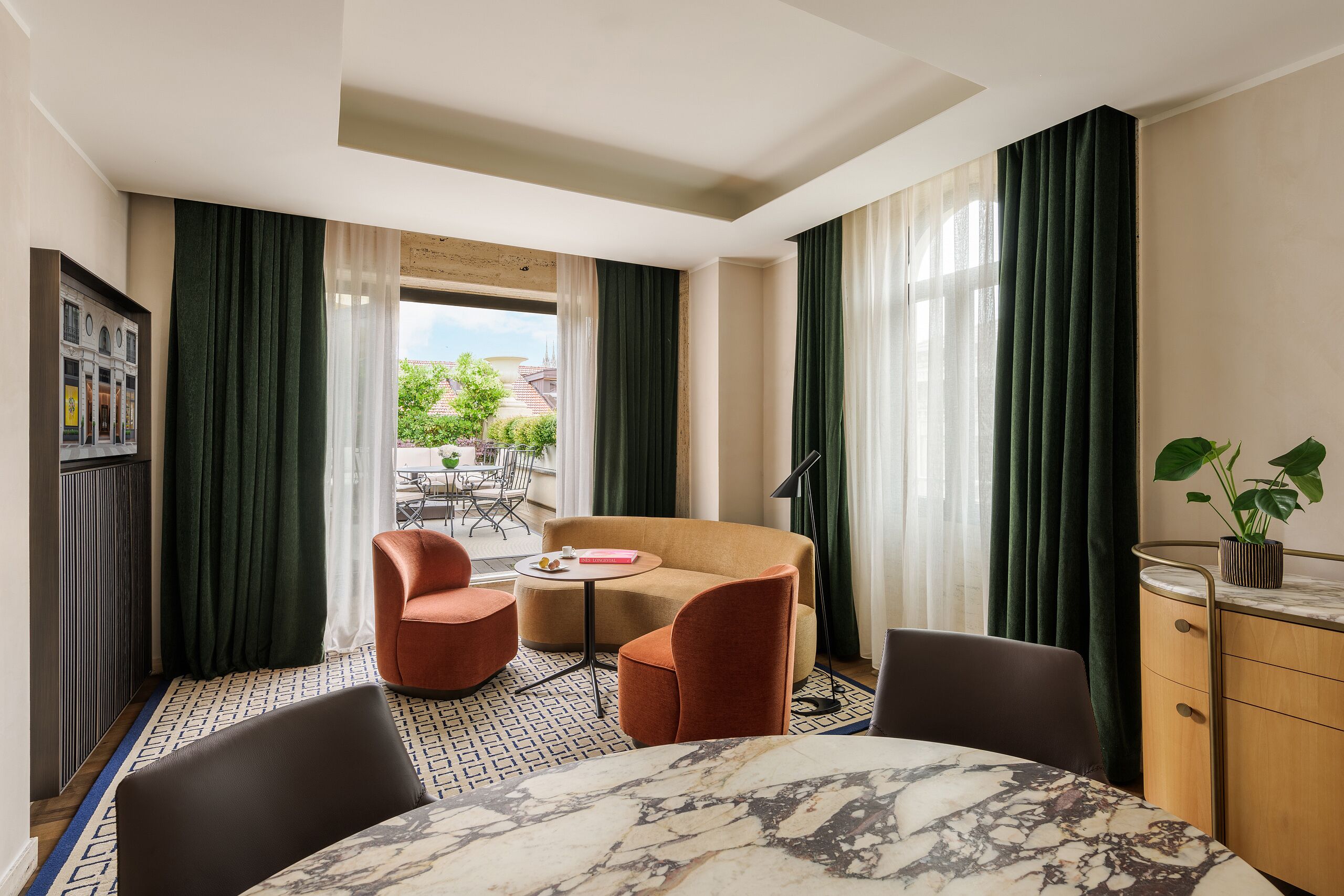 A hotel room with a marble dining table, two armchairs, a sofa, a small round table with a book and a cup, a cabinet with a plant, and a view of the outside through large windows.