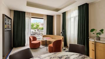 A hotel room with a marble dining table, two armchairs, a sofa, a small round table with a book and a cup, a cabinet with a plant, and a view of the outside through large windows.