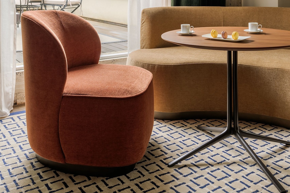 Un hotel moderno con vistas a una catedral, una mesa redonda con dos tazas y macarons, un sofá marrón y una alfombra estampada.