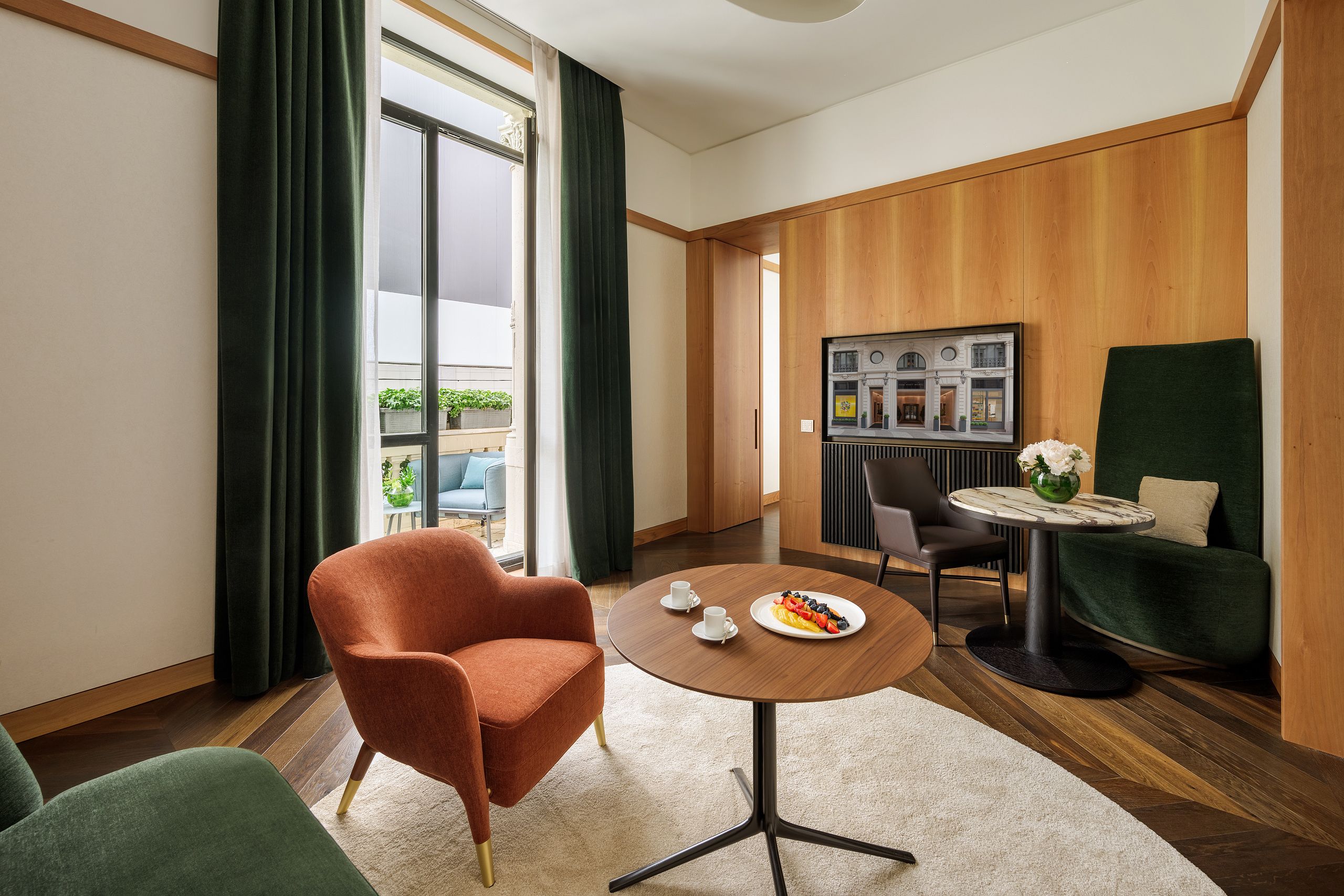 A hotel room with a large window, a round table with a plate of fruit and two cups, a green armchair, and a view of a building through the window.