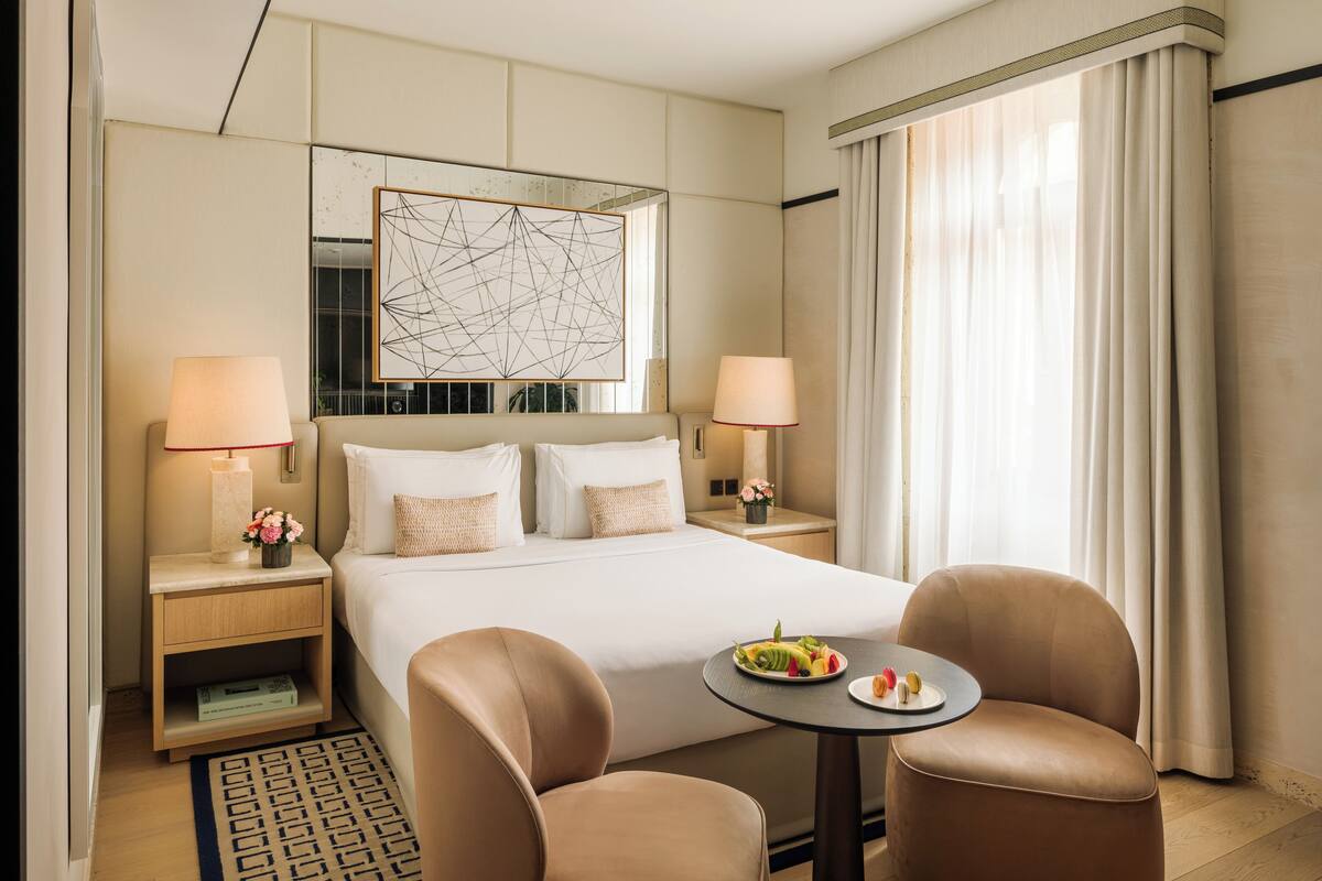 A hotel room with a large bed, two bedside tables with lamps, a small round table with a plate of fruit, and two chairs.