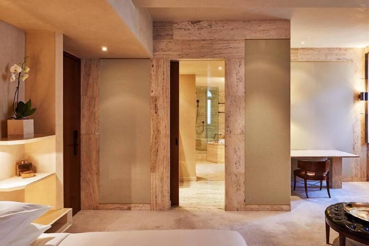 A modern hotel room with a bed, a desk, and a chair. There is a bathroom visible through a glass door.