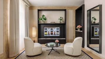Een moderne woonkamer met twee witte fauteuils, een ronde glazen salontafel en een open haard met daarboven een ingelijst beeld. Boven de open haard staan twee potplanten op planken en aan de muur hangt een grote spiegel.