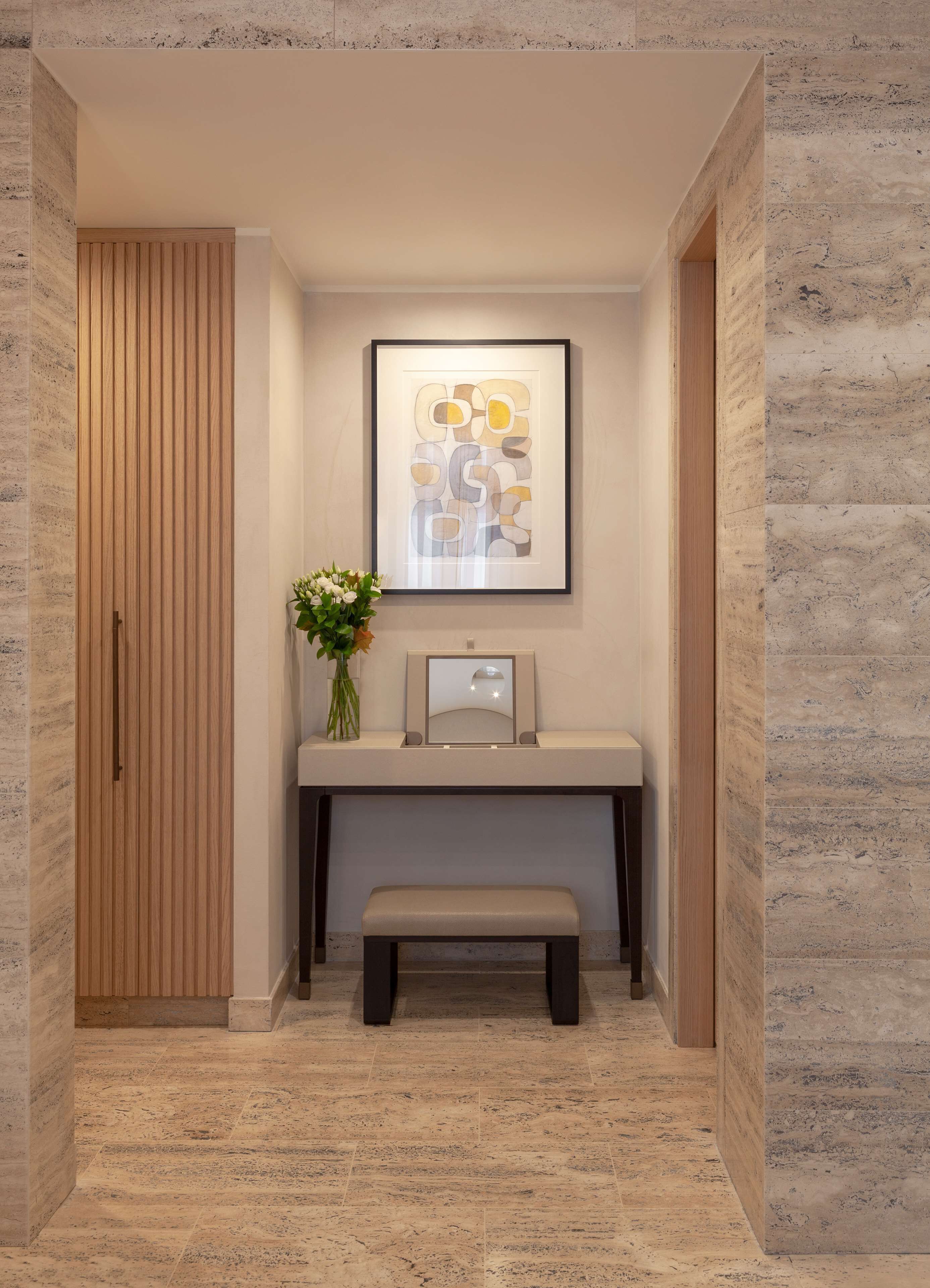 A hallway with a wooden door, a bench, a framed abstract artwork, and a vase with flowers.