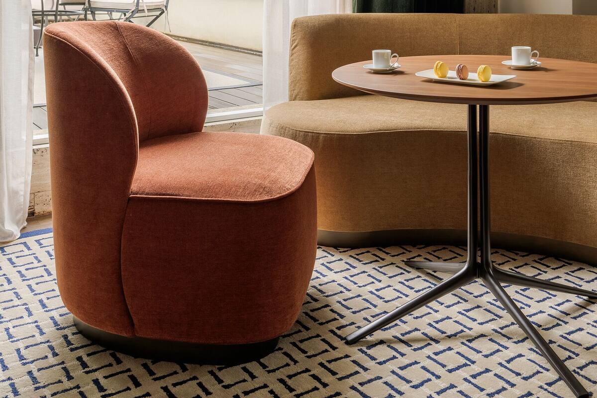 A modern hotel room with a view of a cathedral, a round table with two cups and macarons, a brown sofa, and a patterned rug.