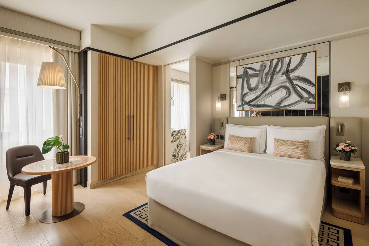 A modern hotel room with a large bed, a round table with a chair, a potted plant, and a large abstract painting on the wall.