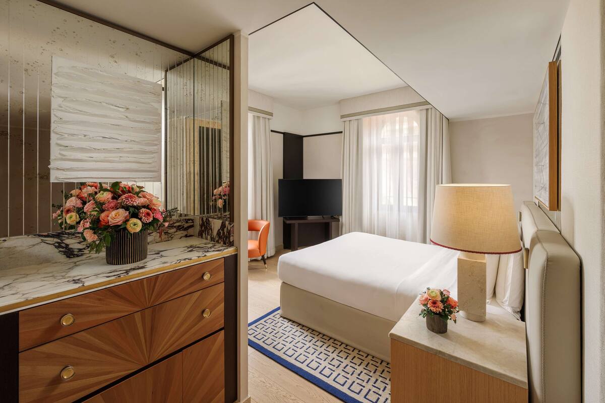 A hotel room with a marble vanity, a bed with a white comforter, a television, and a lamp.