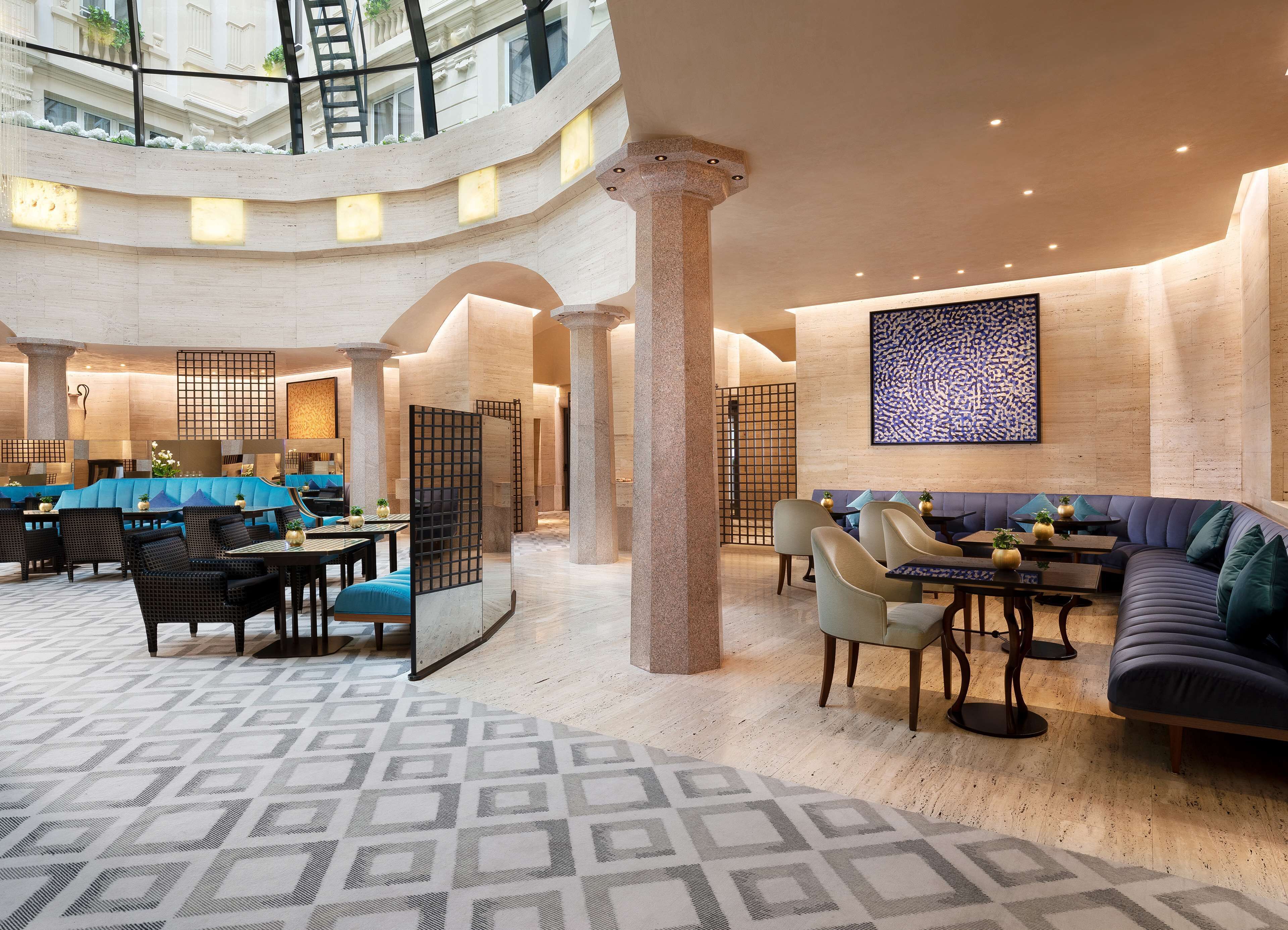 Un hall d’entrée spacieux avec un plafond haut, comprenant un espace de détente central avec des canapés et des tables, le tout entouré de colonnes et agrémenté d’un tapis à motifs.