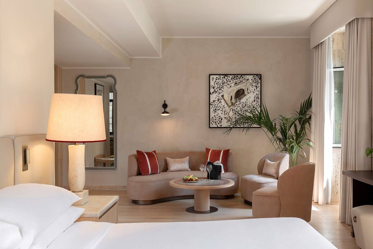 A modern hotel room with a bed, a sofa, a coffee table, and a dining table. There is a large mirror, a wall-mounted lamp, and a potted plant.