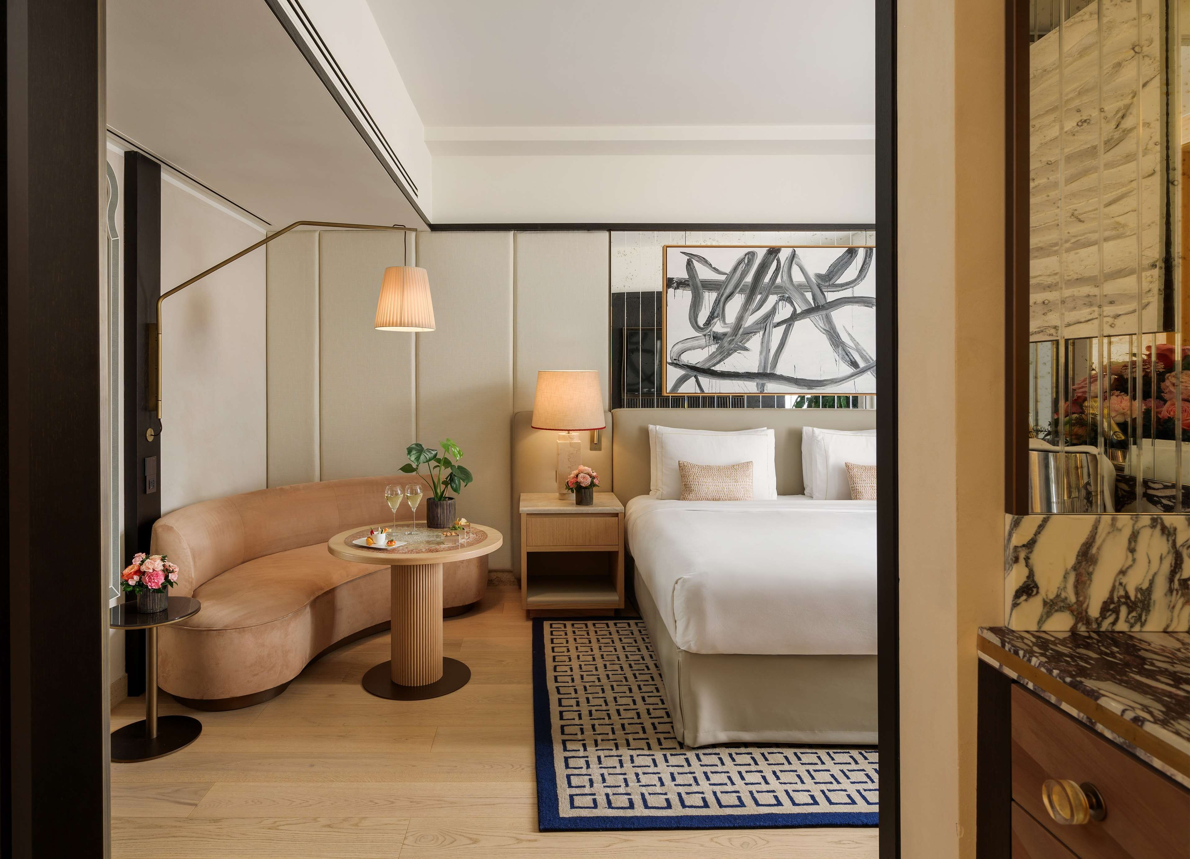 A modern hotel room with a curved sofa, a round table with drinks, a bed with white linens, a nightstand with a lamp, and a large abstract painting on the wall.