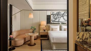 A modern hotel room with a curved sofa, a round table with drinks, a bed with white linens, a nightstand with a lamp, and a large abstract painting on the wall.