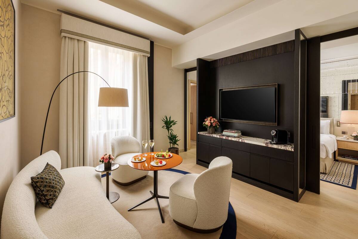 A modern hotel room with a living area featuring a sofa, a round table with a vase of flowers, a flat-screen TV mounted on a dark wall, and a large floor lamp.