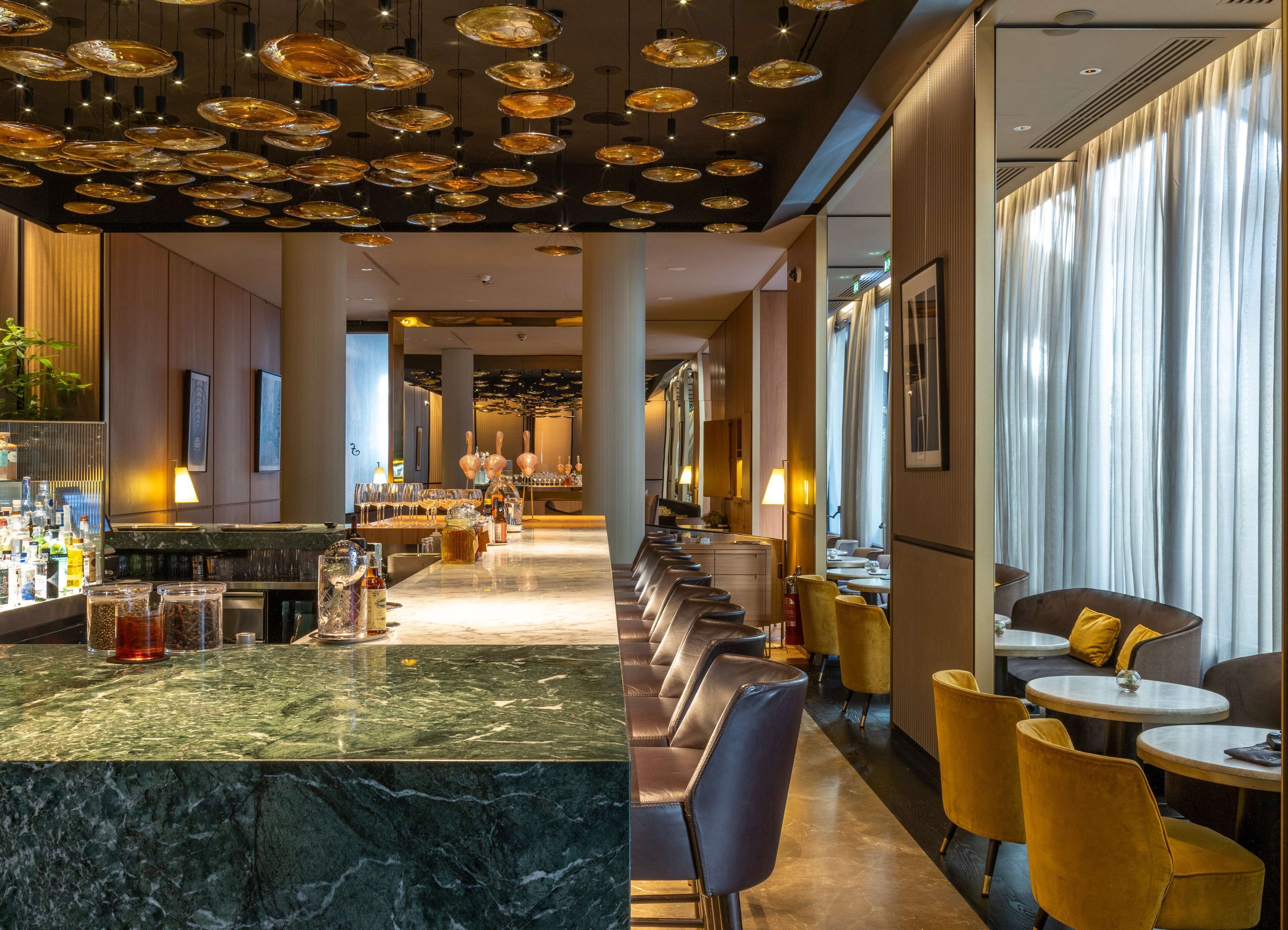 Eine moderne Bar mit Marmorplatte, verschiedenen Sitzmöglichkeiten wie Barhocker und runden Tischen, und einer Decke mit runden Leuchten.