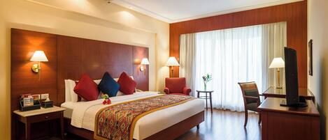 A hotel room with a large bed, two bedside lamps, a small table with a plant, a TV on a stand, and a wooden headboard.