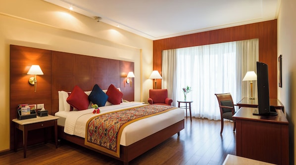 A hotel room with a large bed, two bedside lamps, a small table with a plant, a TV on a stand, and a wooden headboard.