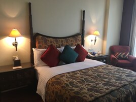 A hotel room with a large bed, two bedside lamps, a nightstand, a chair, and a patterned headboard.