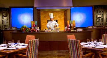 A chef preparing food at a counter in a restaurant with a brick wall backdrop and two large screens on either side.