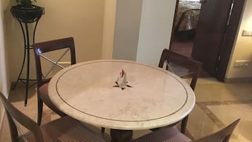 Une table de salle à manger ronde, entourée de quatre chaises, un pot de fleurs et, à travers une porte ouverte, on aperçoit une chambre.