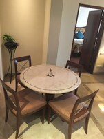 A round dining table with four chairs, a potted plant, and a view of a bedroom through an open door.