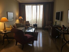 Una sala de estar bien iluminada con sofá, sillón y una mesa de centro. Hay un televisor montado en la pared, una lámpara en una mesita auxiliar y una ventana con cortinas.