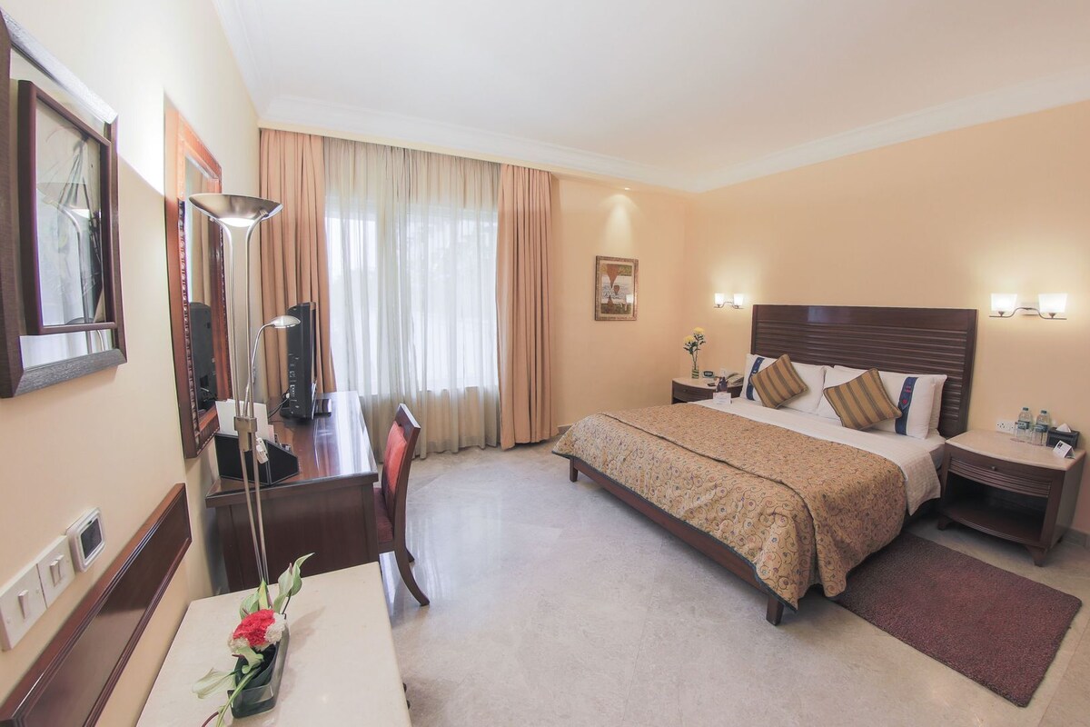 A hotel room with a bed, desk, chair, lamp, bedside table, and a vase with flowers.