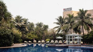 Ein Schwimmbecken mit Liegestühlen und einem Pavillon, umgeben von Palmen und einem Gebäude mit der Aufschrift 'The Lalit Residency'.