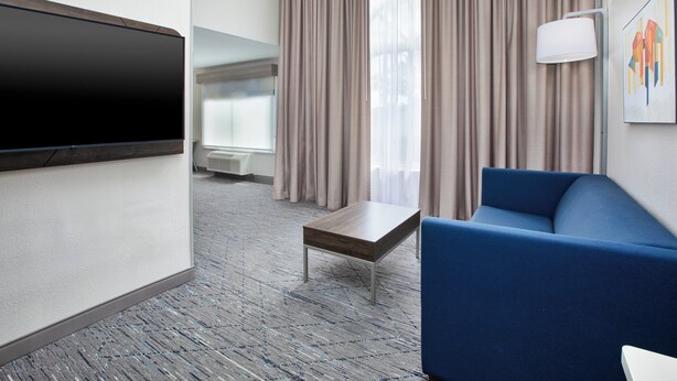 A hotel room with a flat-screen TV, a blue sofa, a small wooden table, and a large window with curtains. - Holiday Inn Express & Suites W. Monroe by IHG (West Monroe)