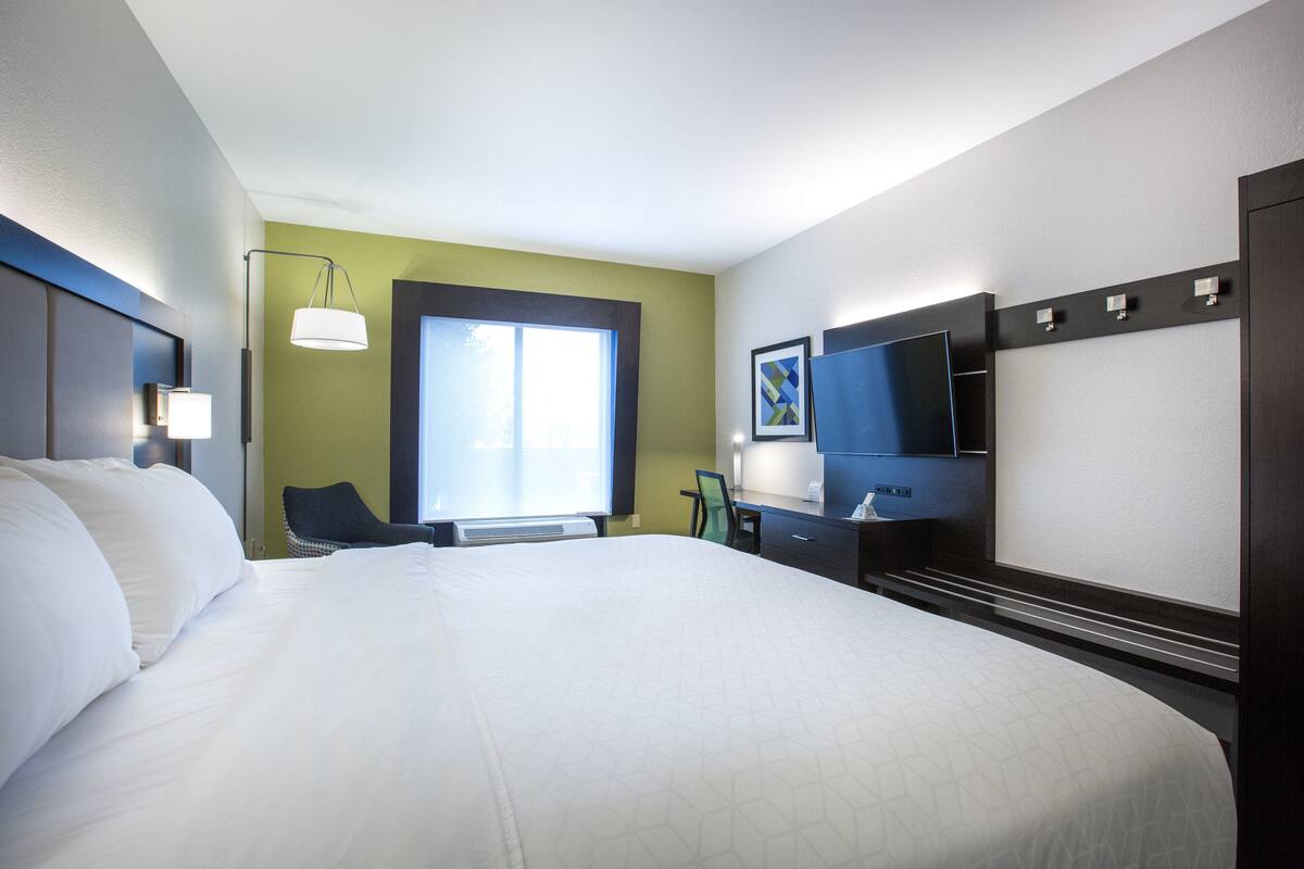 A hotel room with a large bed, a desk with a flat-screen TV, a chair, and a window with blinds.