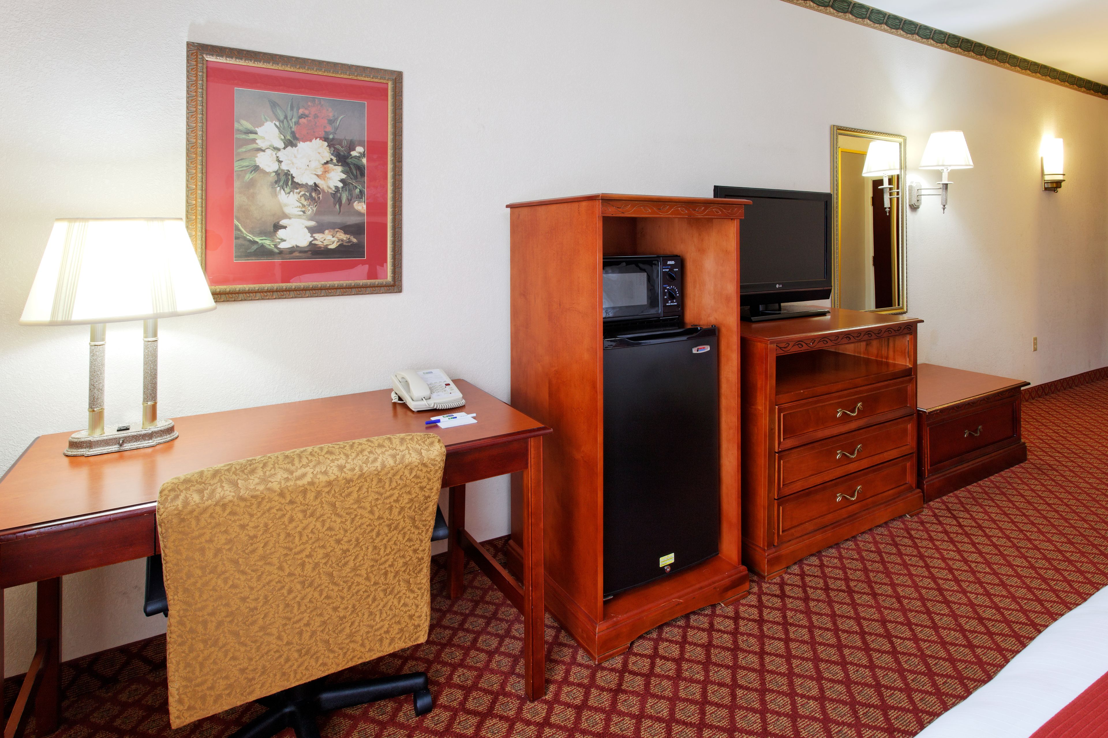 A hotel room with a wooden desk, a chair, a microwave, a refrigerator, a telephone, a television, and a mirror with wall sconces.