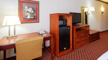 A hotel room with a wooden desk, a chair, a microwave, a refrigerator, a telephone, a television, and a mirror with wall sconces.