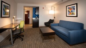 A hotel room with a blue sofa, a desk with a chair, a coffee table, and a kitchenette in the background.