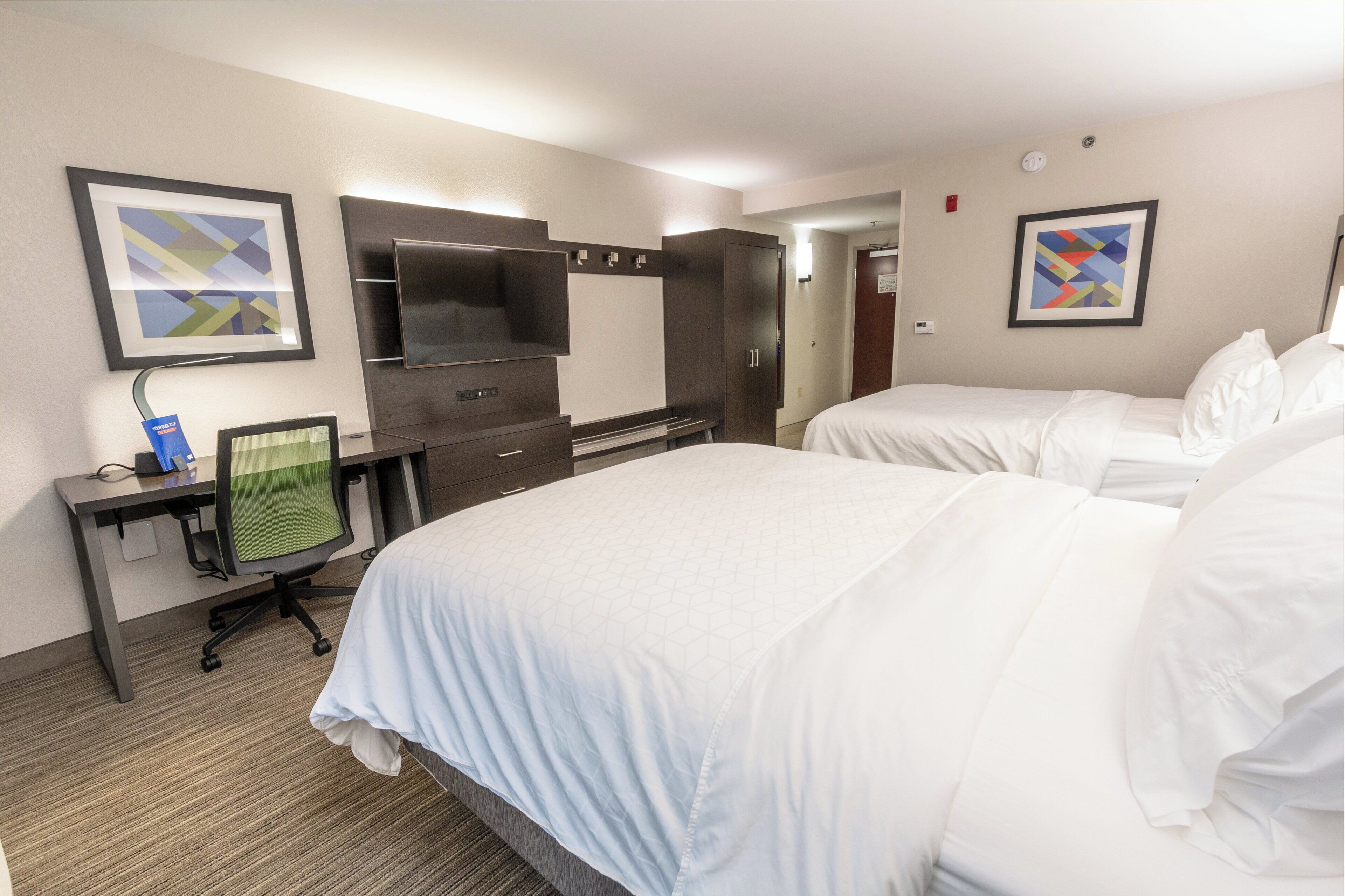 Habitación de hotel con dos camas, un escritorio con computadora, un televisor y obras de arte en las paredes.