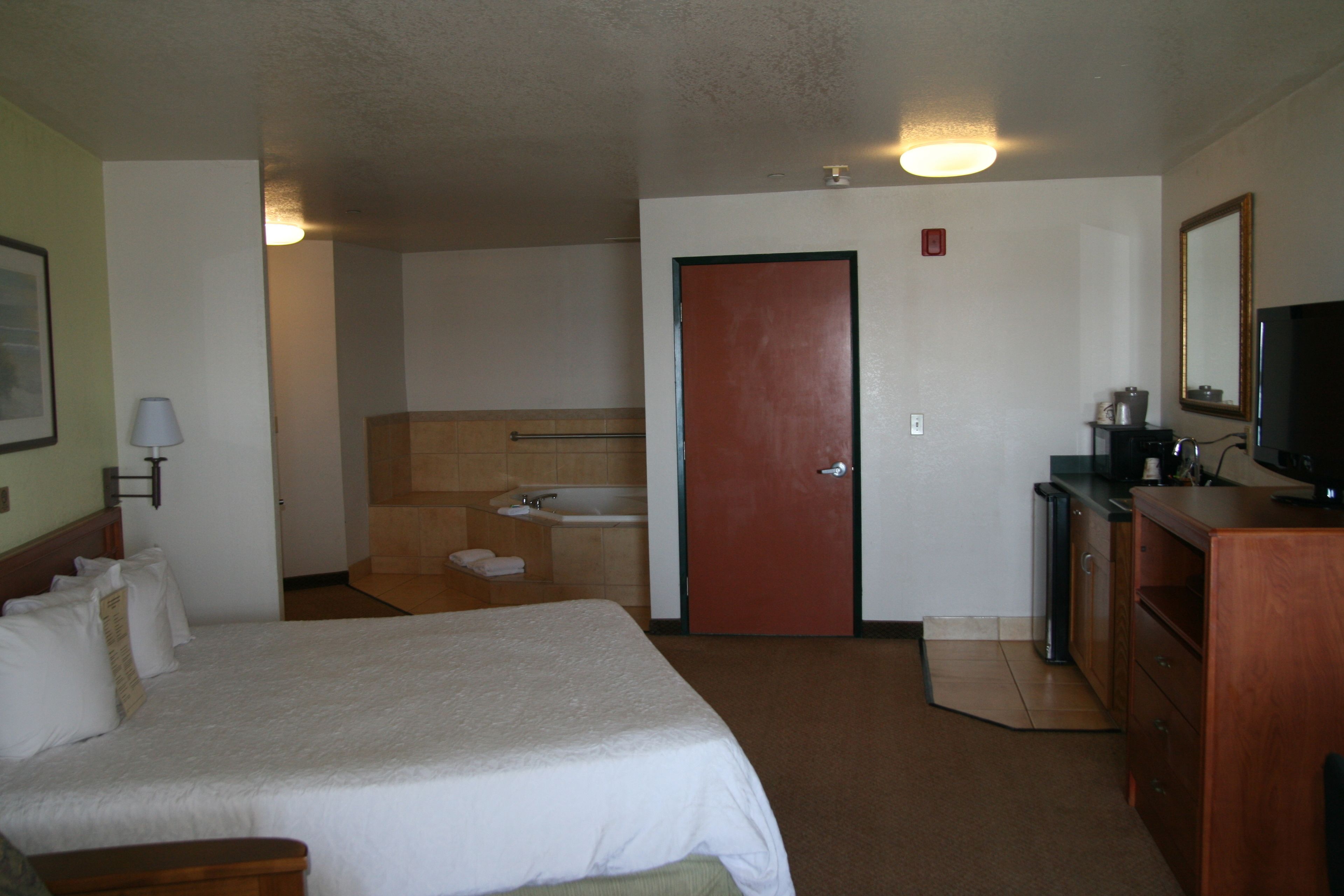 A hotel room with a bed, a desk, a television, a mirror, a door, and a bathroom with a sink and a bathtub.