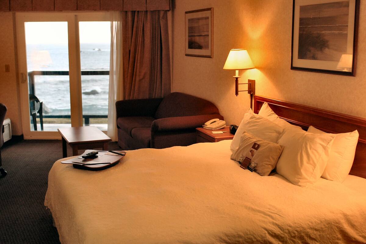 A hotel room with a bed, a desk, a chair, a lamp, and a view of the ocean.