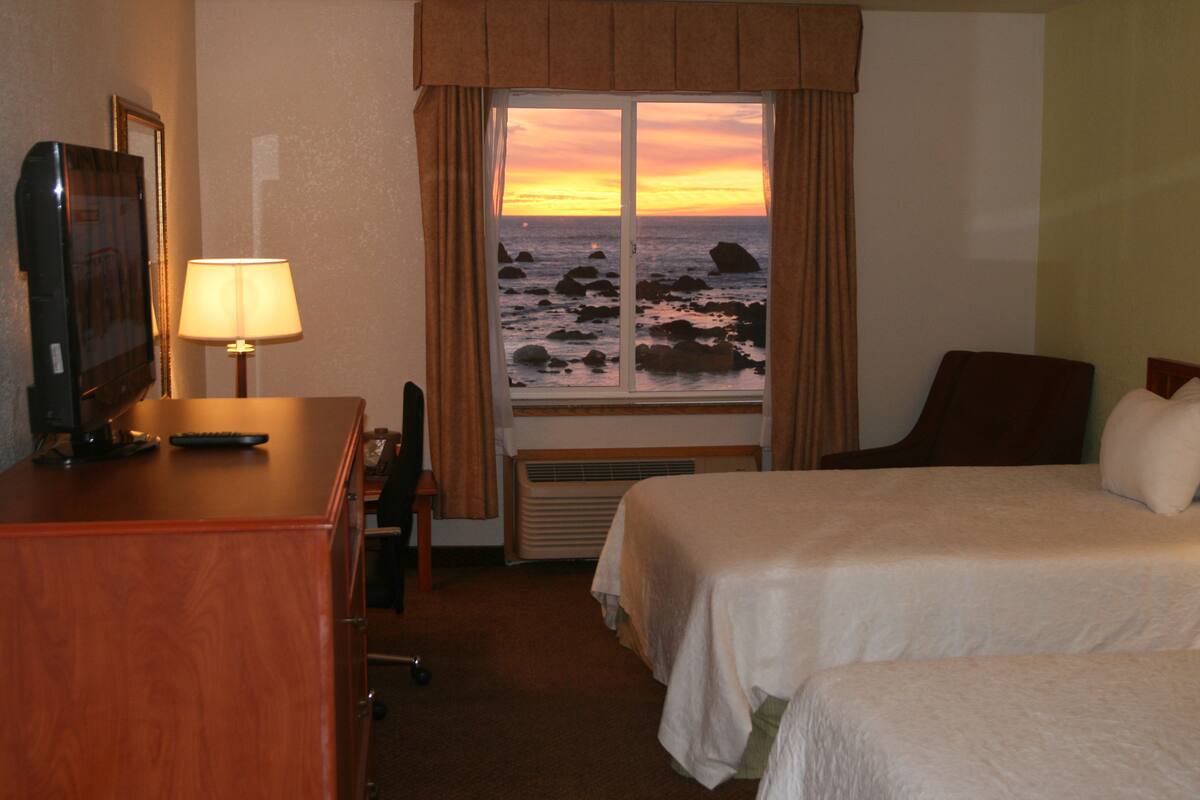A hotel room with a bed, a desk with a lamp, a chair, and a view of the ocean at sunset.