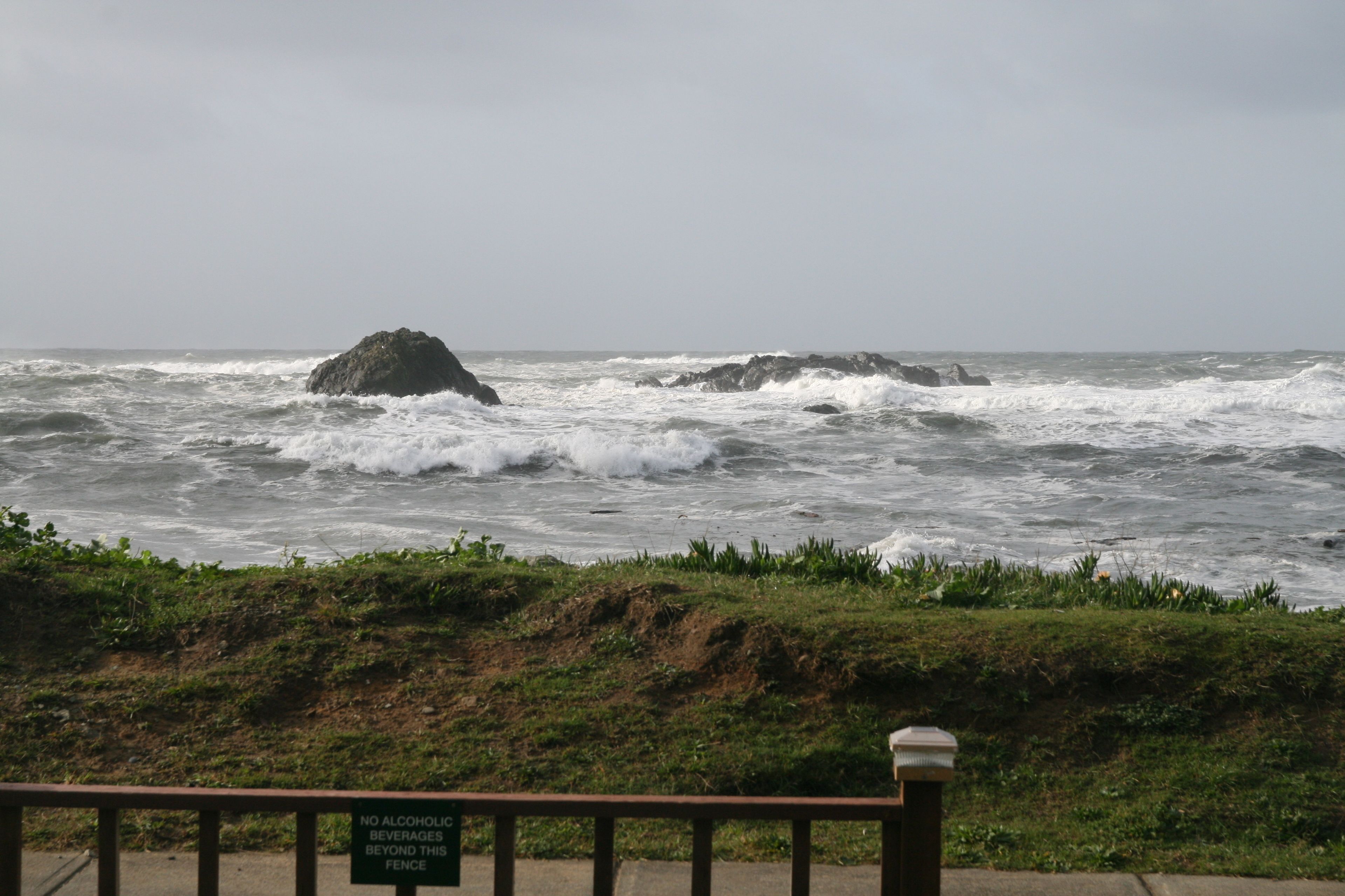 A coastal view with rough sea waves and a rocky outcrop. A sign on the fence reads "No alcoholic beverages beyond this fence."