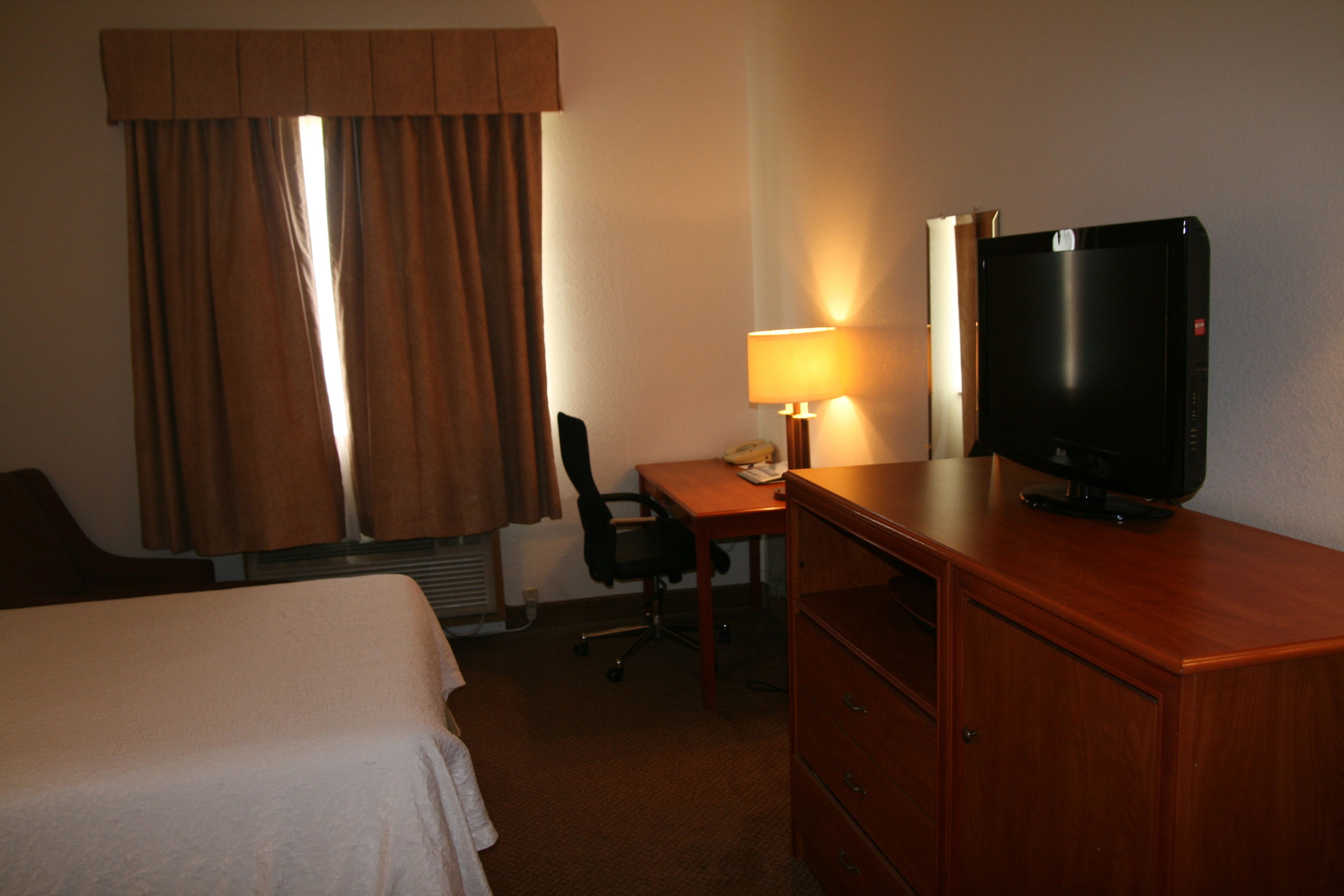 A hotel room with a bed, a desk with a lamp, a television, and a chair.