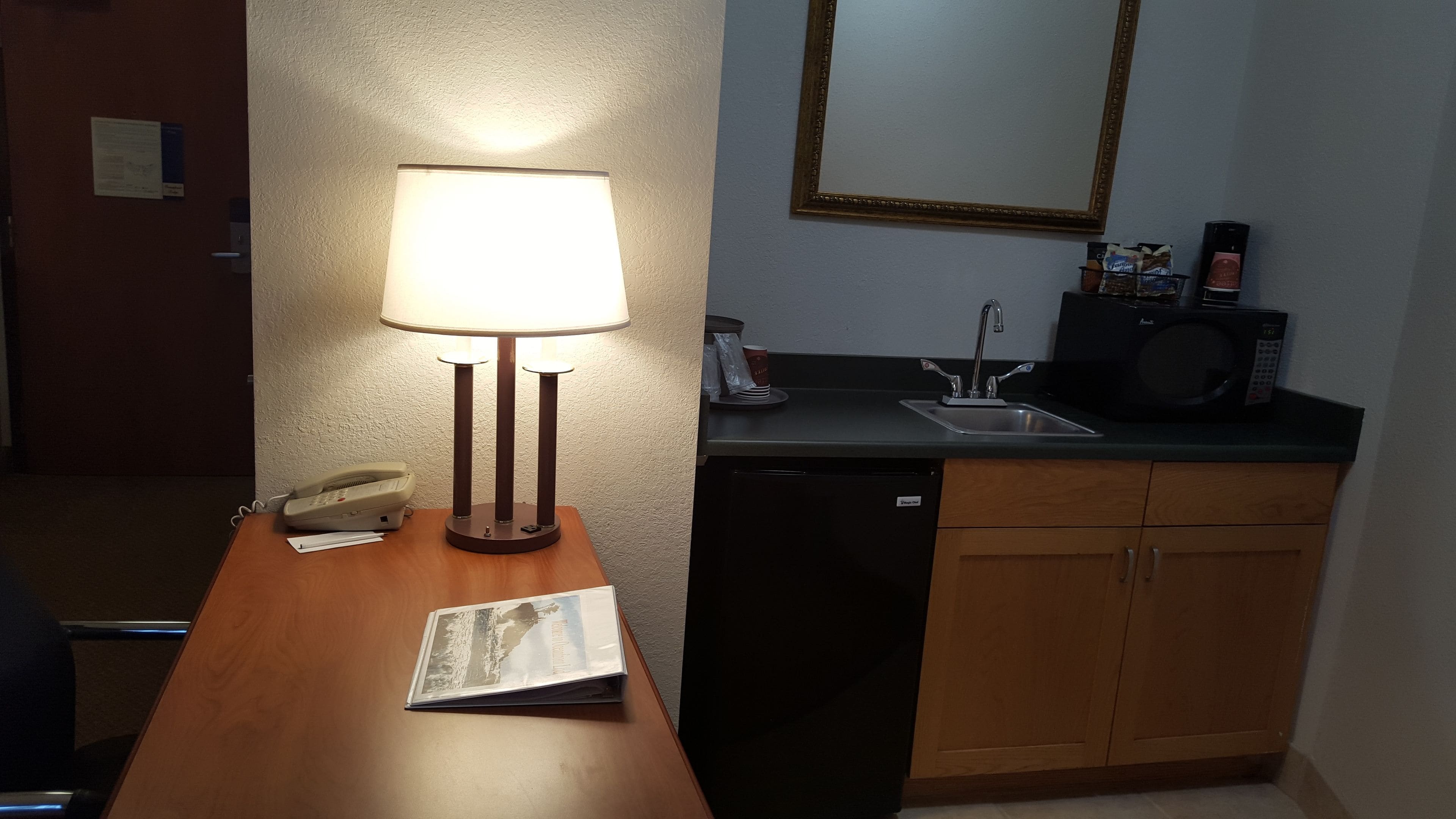 A hotel room with a wooden desk, a lamp, a telephone, a magazine, a sink, a microwave, and wooden cabinets.