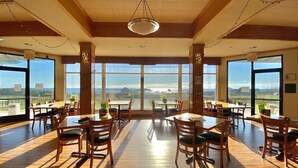 A spacious dining area with large windows offering a view of the sea and landscape. Tables and chairs are arranged for dining.